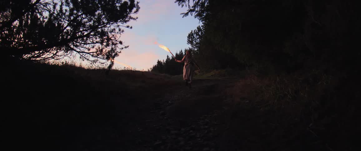 A mysterious woman holding a fiery torch walks up a dark hill through a forest at twilight in slow motion