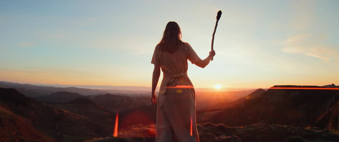 A woman in a long dress stands on a mountain peak at sunset, holding a flaming torch high against the epic landscape
