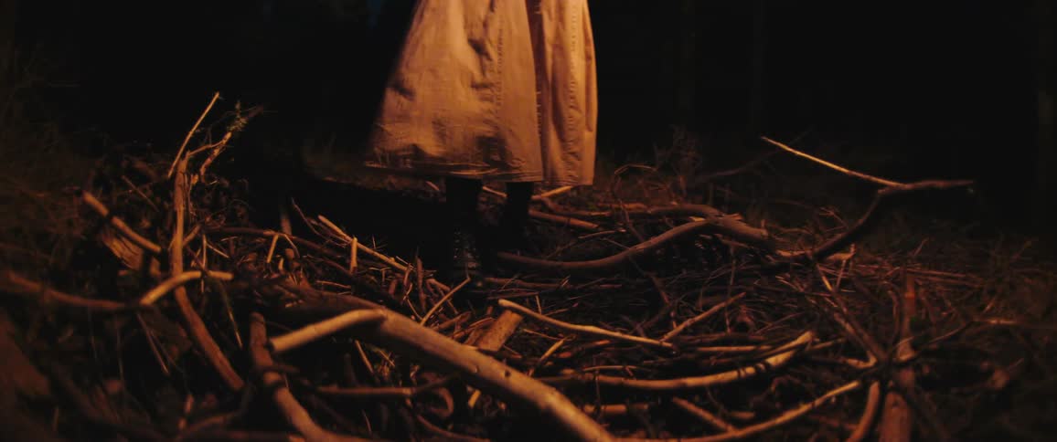 A figure in a long trench coat walks in slow motion through a dark, eerie forest at night, stepping over a bed of broken sticks