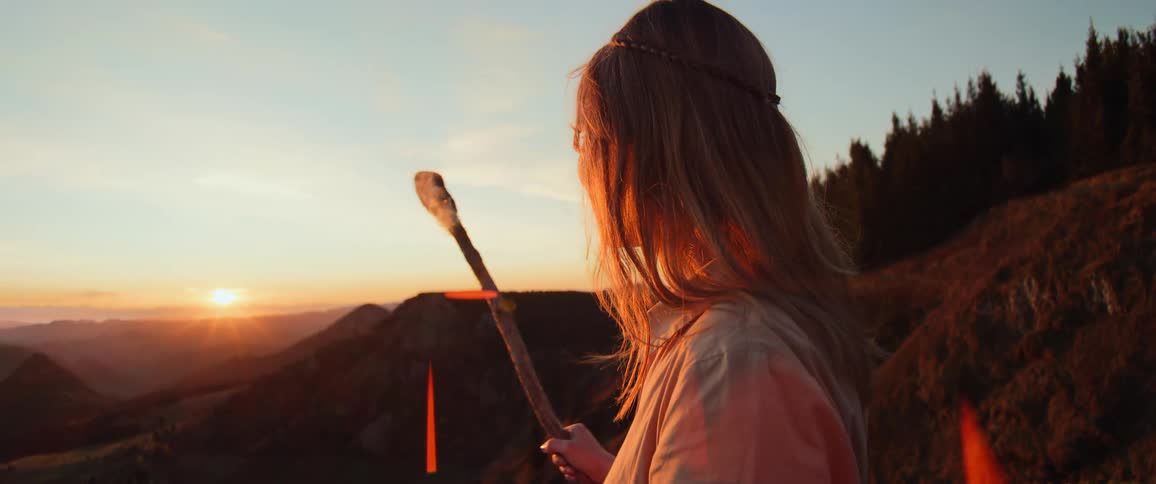 A beautiful young woman with a staff stands on a mountaintop, peacefully watching the golden sunset over the rolling hills