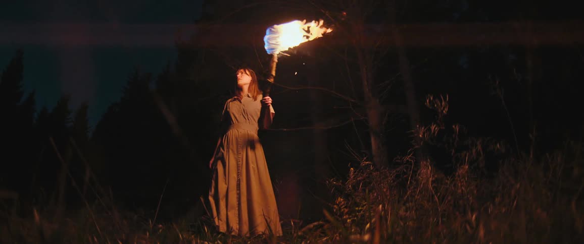 A woman in a vintage dress holding a flaming torch stands in a dark forest at night before walking away into the darkness