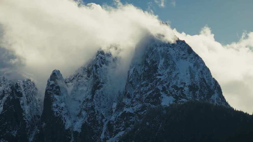 A majestic snow-covered mountain peak is shrouded in slow-moving clouds against a blue sky
