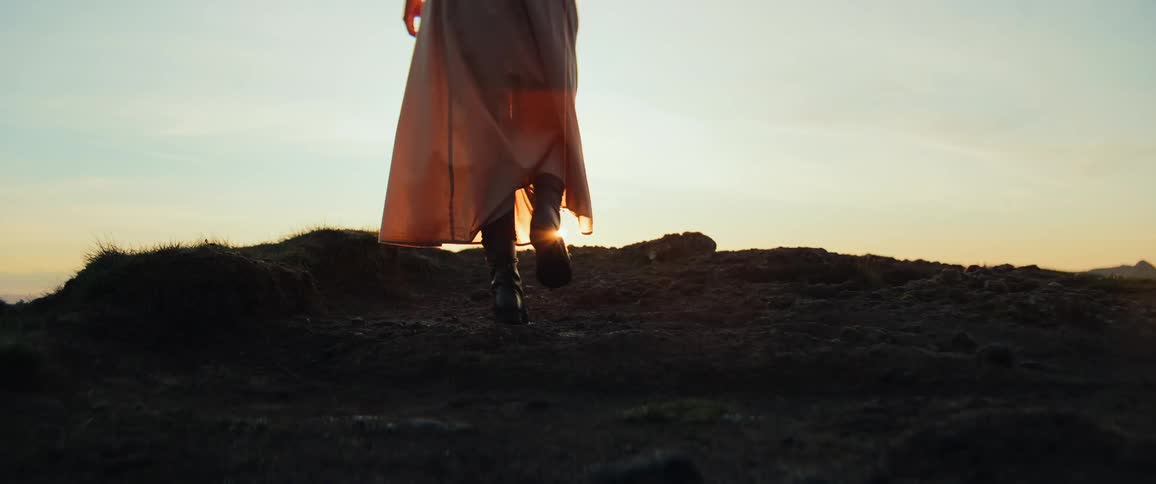 A woman's boots walk over a silhouetted hill towards a beautiful sunset in a dramatic slow-motion shot with golden lens flare