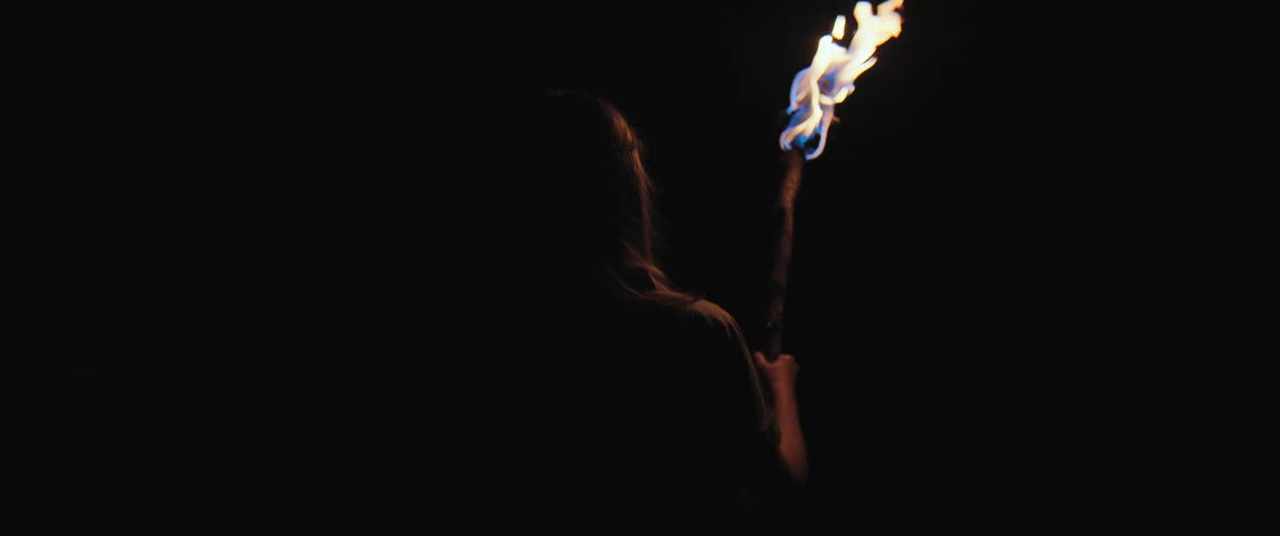 A silhouetted figure holds a burning torch high, its flame flickering in slow motion against a pitch-black background