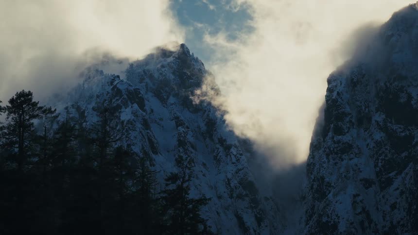 A slow dolly zoom reveals majestic snow-covered mountain peaks shrouded in dramatic, sunlit clouds