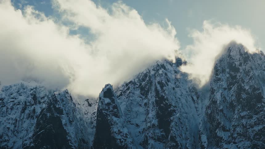 A cinematic zoom out from majestic, snow-covered mountain peaks shrouded in swirling white clouds to reveal a vast evergreen forest below