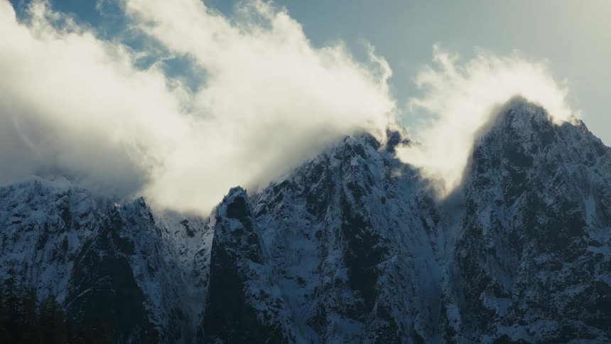 A slow zoom out reveals majestic, snow-covered mountain peaks shrouded in ethereal clouds under a blue sky