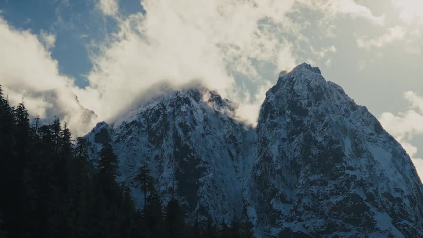 A majestic slow zoom towards snow-covered mountain peaks as clouds drift across a sunny sky