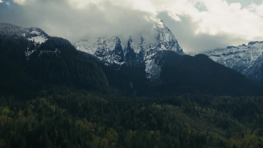 Majestic snow-capped mountain peaks emerge from swirling clouds above a dense evergreen forest