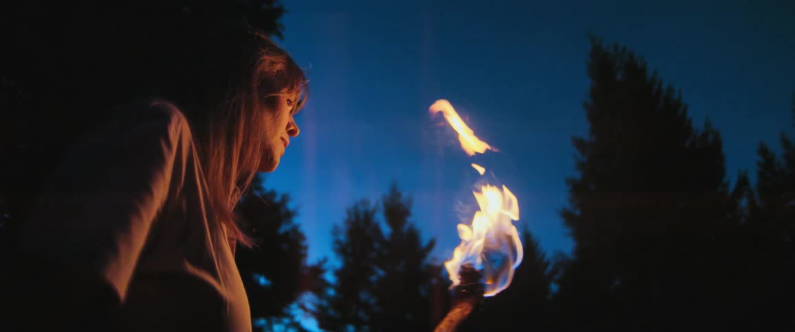 A young woman watches a burning torch extinguish against the dark blue night sky in a forest