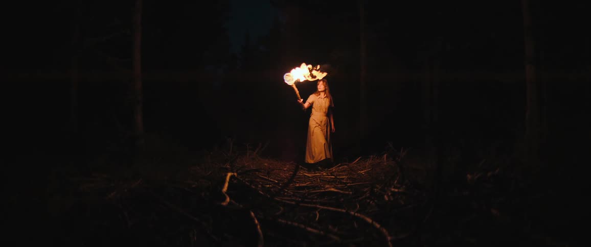 A woman in a vintage dress walks through a dark, spooky forest at night holding a flaming torch