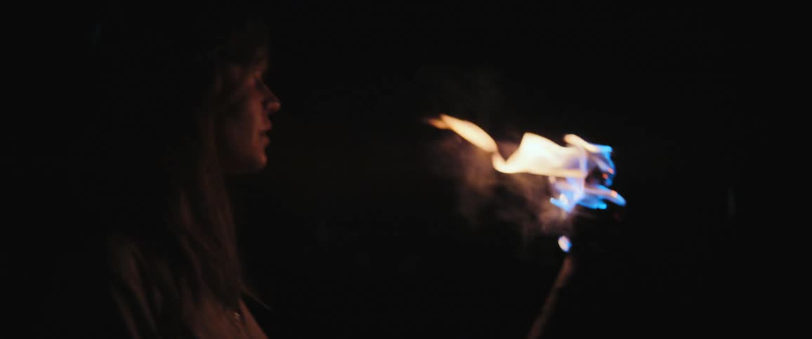 A woman's face is illuminated by a flickering torch as she cautiously explores the darkness at night