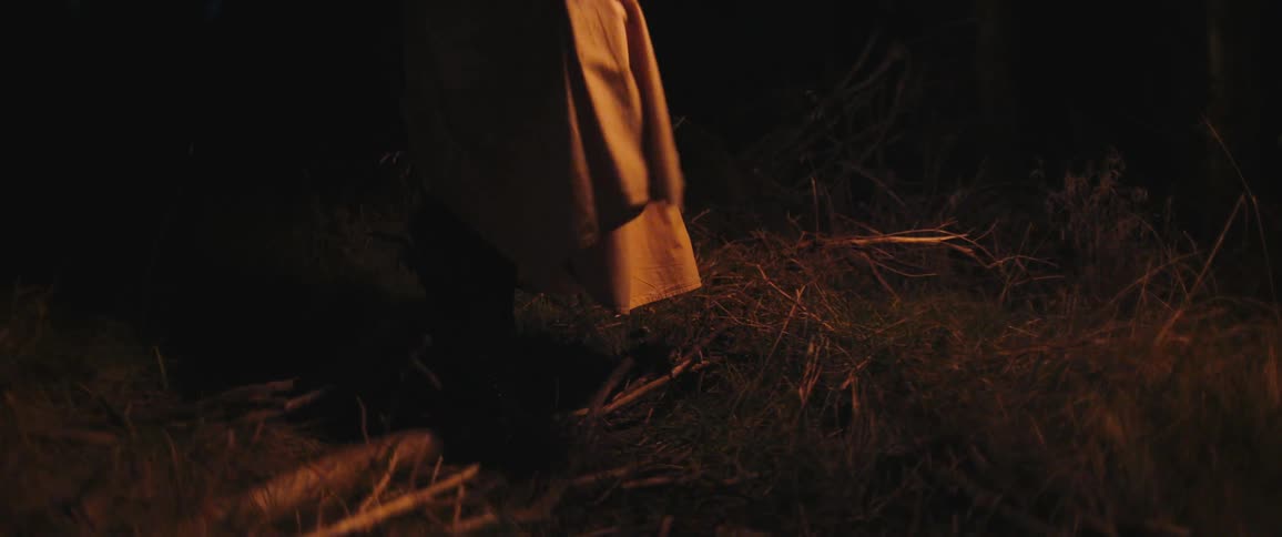 A person wearing a long coat and boots shuffles their feet in a dark forest at night, lit by a warm, mysterious glow