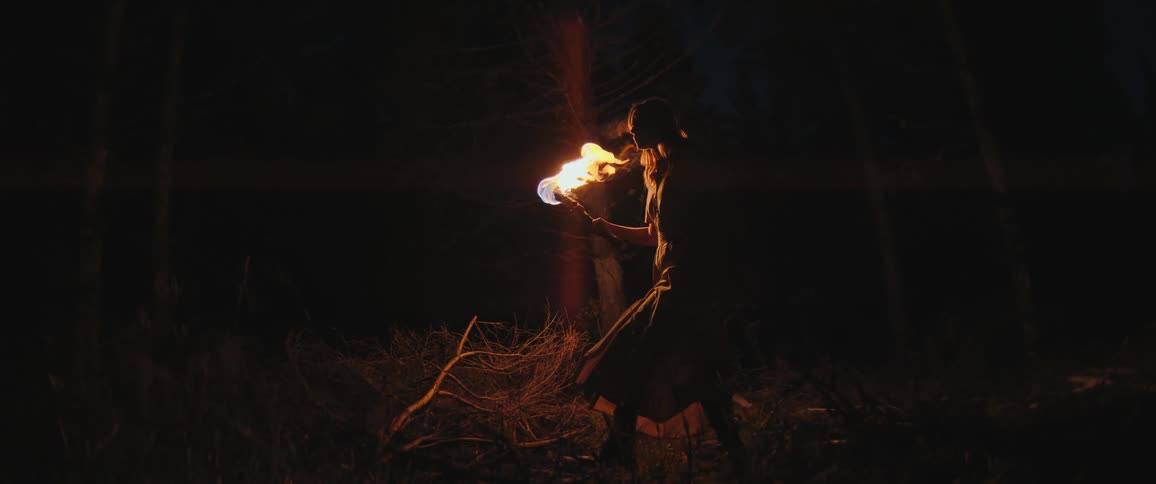 A woman walks slowly through a dark forest at night, illuminating her path with a large, flaming torch in slow motion