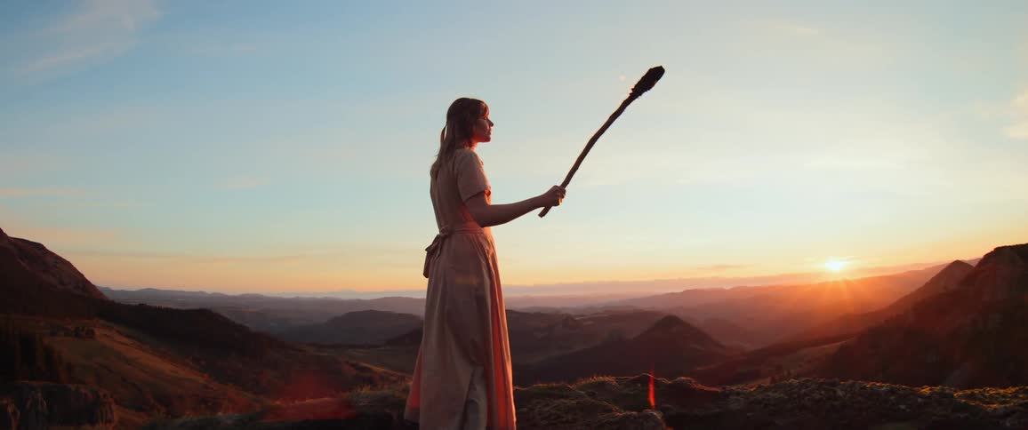 A woman in a flowing dress holds a flaming torch on a mountaintop, overlooking a beautiful landscape at sunset