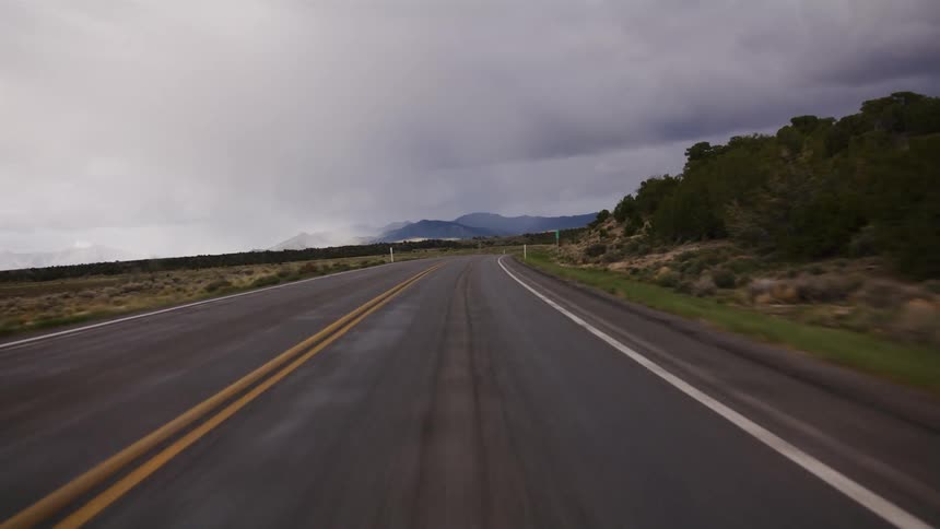 A point-of-view shot driving on a wet, two-lane rural highway through an arid landscape under a dark and dramatic cloudy sky