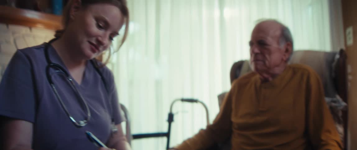 A female nurse in scrubs smiles while taking notes during a home visit with an elderly male patient