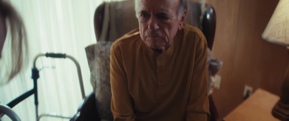 A healthcare worker fills out a medical intake form for a sad, elderly man sitting in a chair in his room