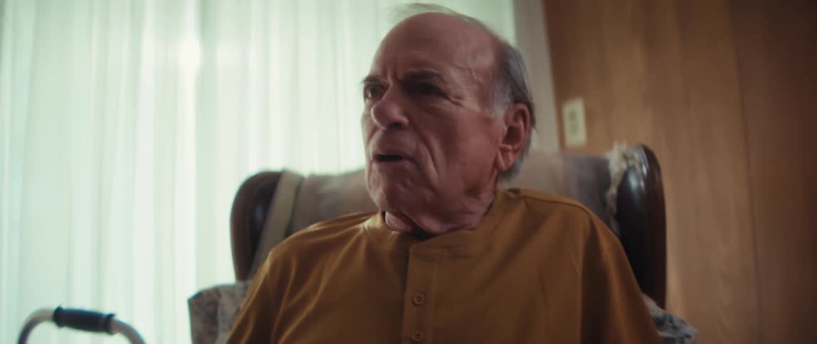 A smiling elderly woman, a pensive elderly man, and an attentive female caregiver have a conversation in a cozy home