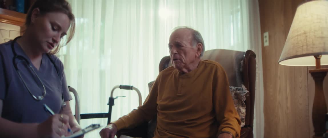 A home healthcare provider consults with a senior man sitting in his armchair next to his walker