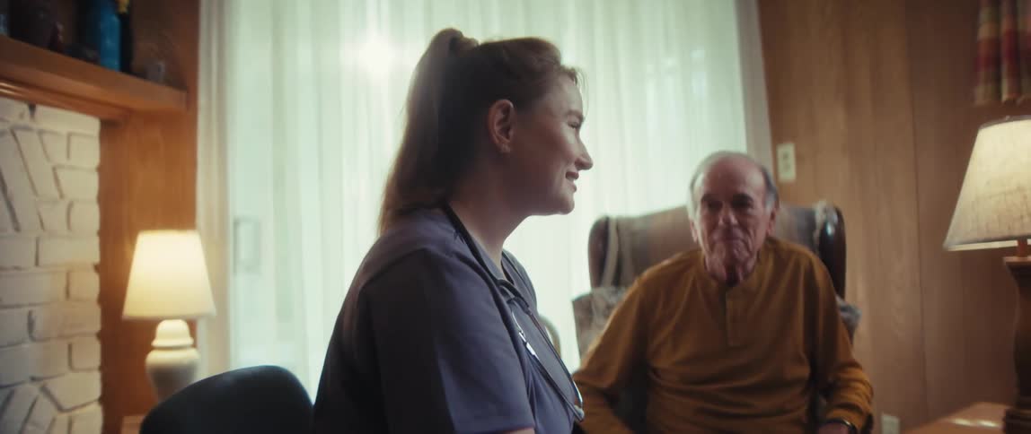 A compassionate nurse provides in-home care for an elderly man, smiling and taking notes during a consultation in his living room
