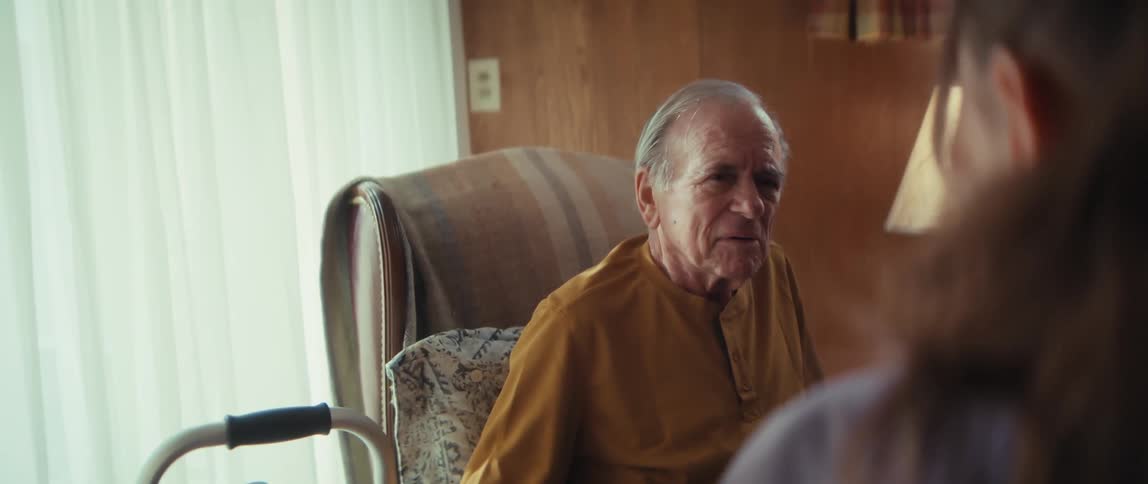 A female home healthcare nurse in scrubs takes notes on a clipboard while having a conversation with an elderly male patient in his living room