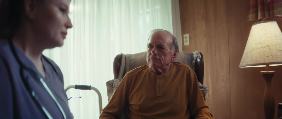 An elderly man listens intently as his wife talks to a home healthcare nurse during a consultation in their living room