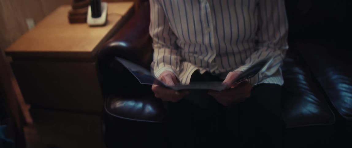 A sad elderly woman sits alone on a couch in a dimly lit room and reads a healthcare brochure