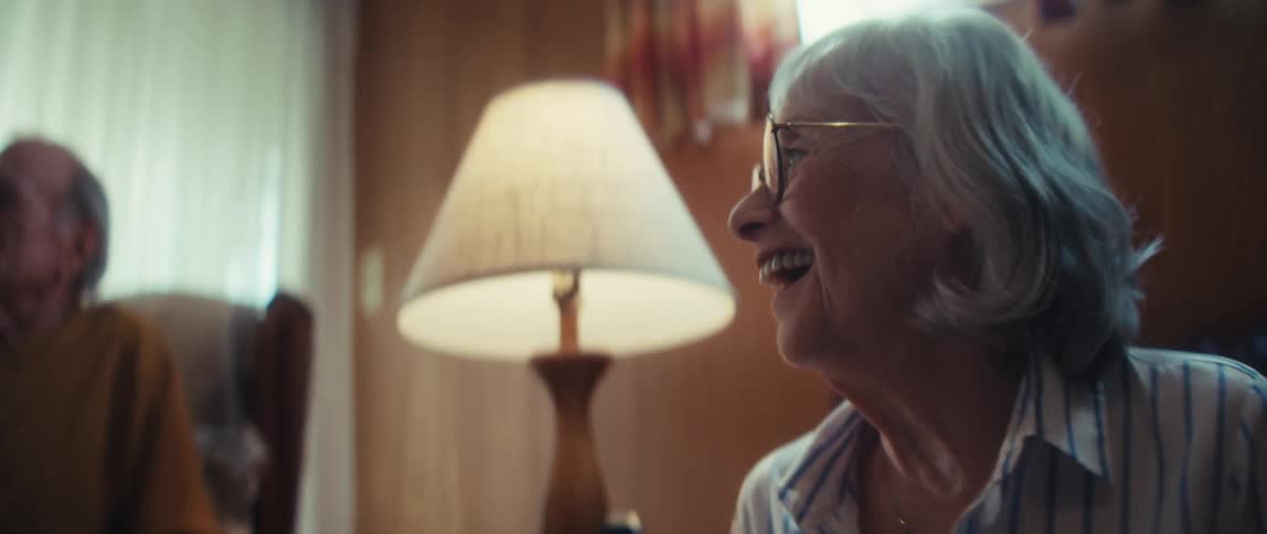 An elderly woman in glasses smiles and laughs while a nurse provides in-home care