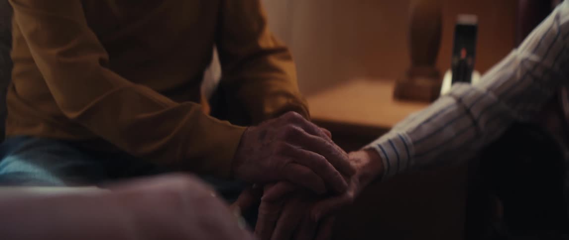 An elderly man comforts his wife, gently holding her hand as they share a warm and loving conversation