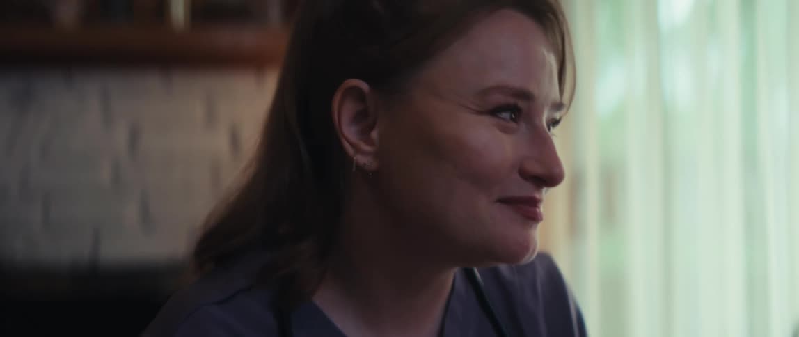 A compassionate female healthcare professional in scrubs smiles warmly and listens with empathy in a softly lit room