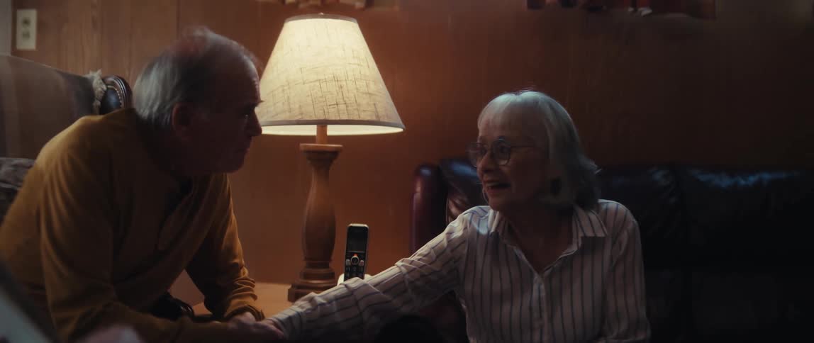 A smiling caregiver looks on as an elderly man and woman share a tender, emotional moment holding hands in a warmly lit room
