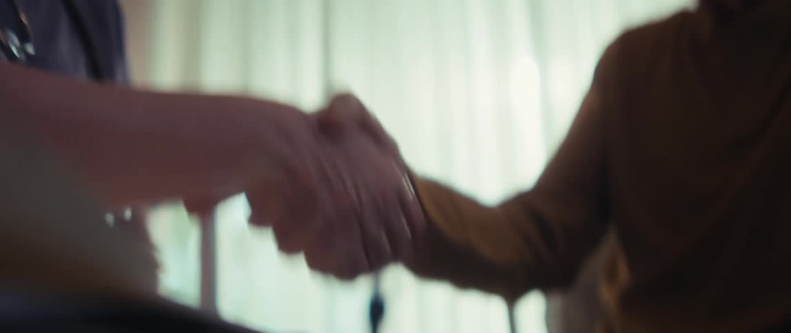 A doctor in scrubs shakes the hand of her elderly patient in a sunlit room in a close-up shot