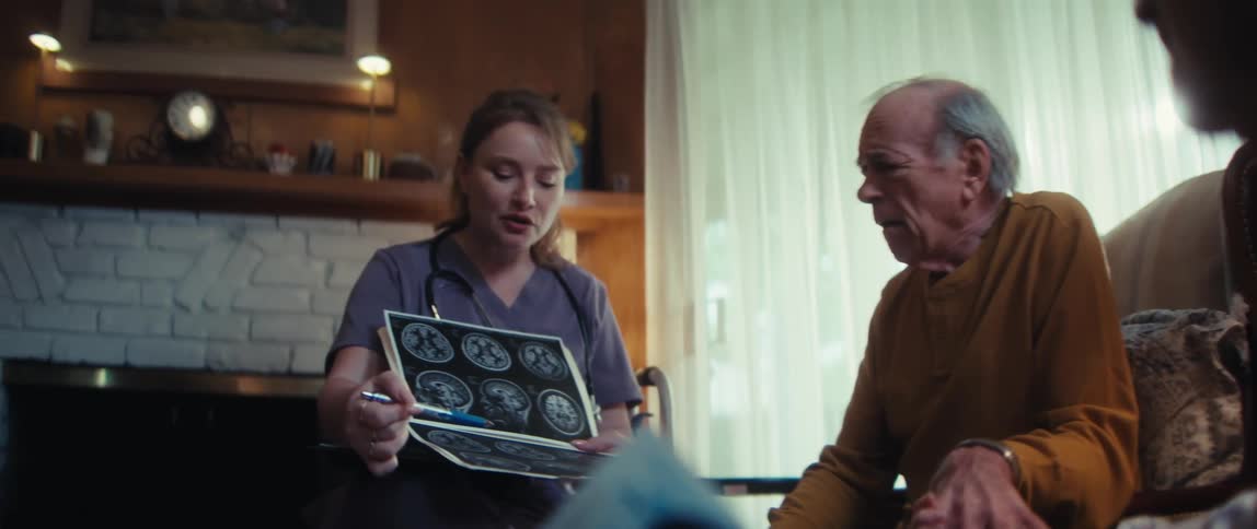 A compassionate home care nurse explains a brain scan diagnosis to a concerned elderly couple holding hands in their living room