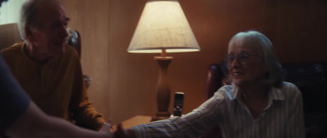 An elderly couple has a warm and serious conversation with a visitor, shaking hands in slow motion in a cozy, lamp-lit room
