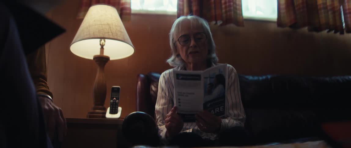 An elderly woman with glasses reads a healthcare brochure while sitting on a couch and talking to someone off-screen