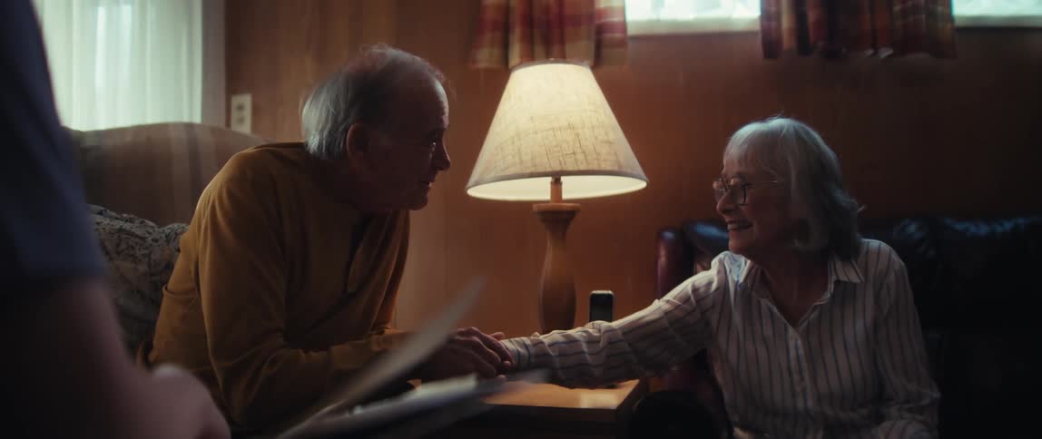 A loving elderly couple holds hands and reminisces, smiling and laughing together in a cozy, lamp-lit living room