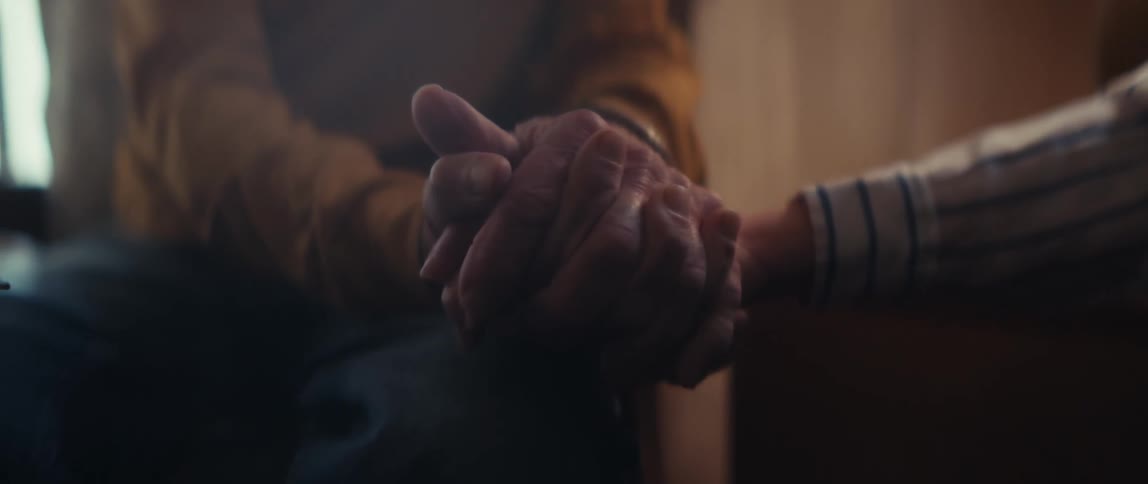 An elderly couple laughs and smiles together in their home, and the camera pans to a close-up of them holding hands