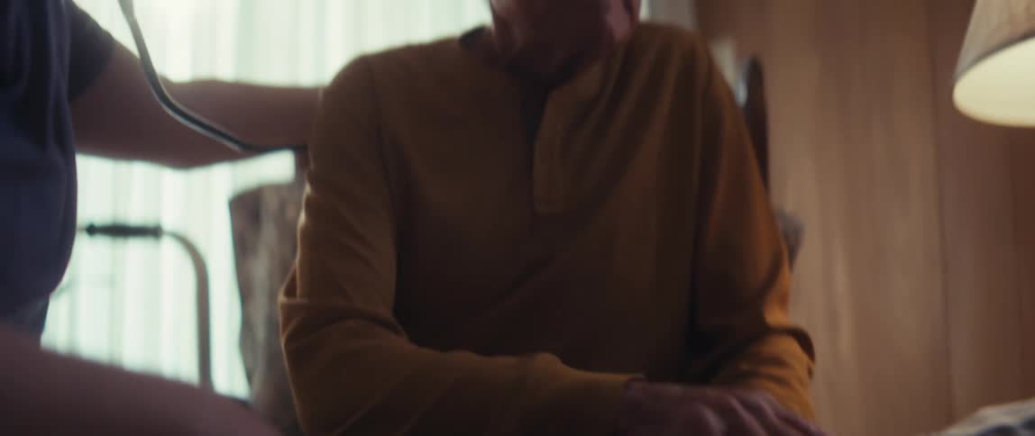 Close up of an elderly woman holding her husband's hand for comfort as a healthcare professional attends to him in their home