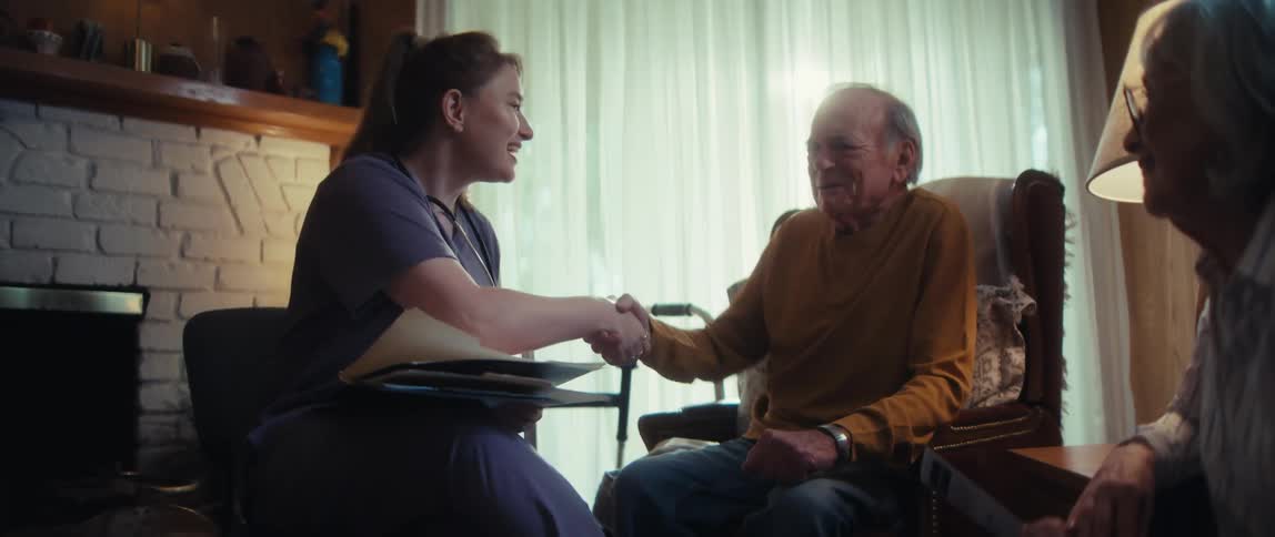A friendly home care nurse explains brain scan results to an elderly couple and offers a reassuring handshake in their sunlit living room