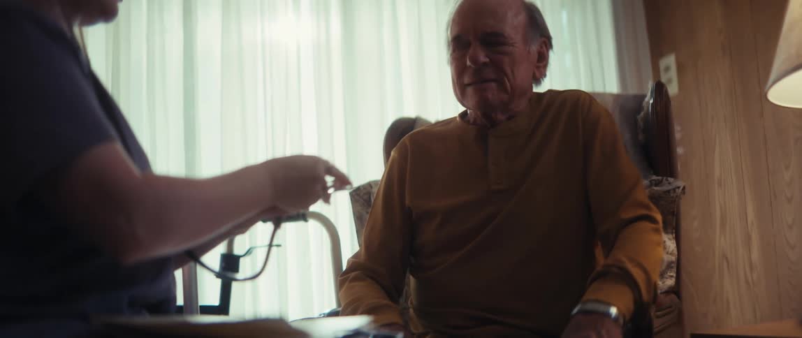 An elderly man receives a medical check-up from a home healthcare nurse who is listening to his chest with a stethoscope