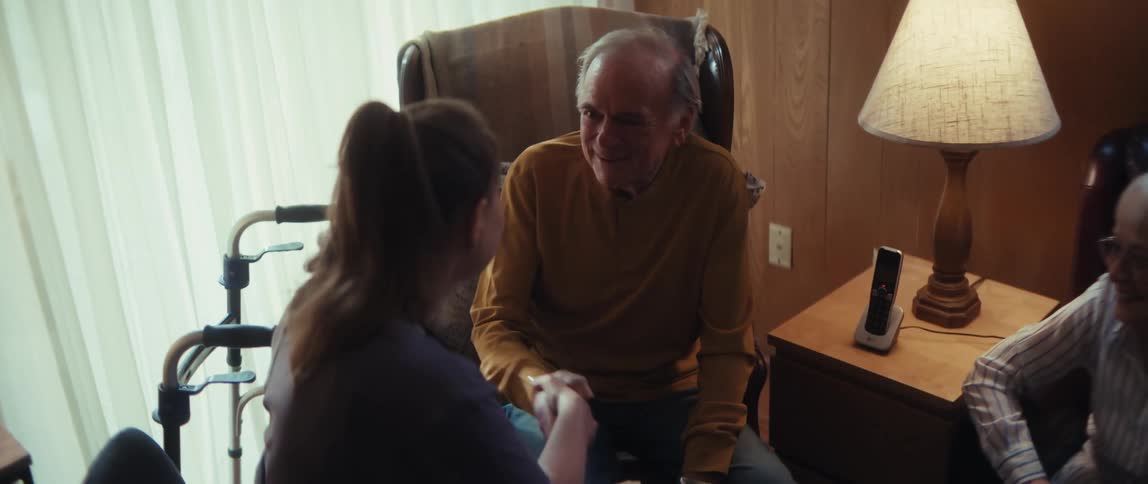 A caregiver offers comfort and support, holding the hands of a smiling elderly couple in their home