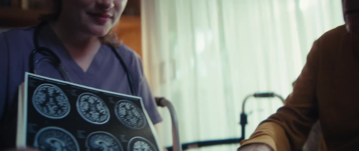A compassionate female nurse discusses a serious diagnosis with a concerned elderly couple, pointing to a brain scan during a home visit