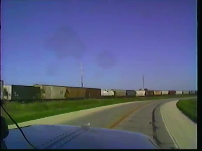 Vintage VHS footage from inside a truck cab shows a long freight train moving alongside a winding rural road on a clear day