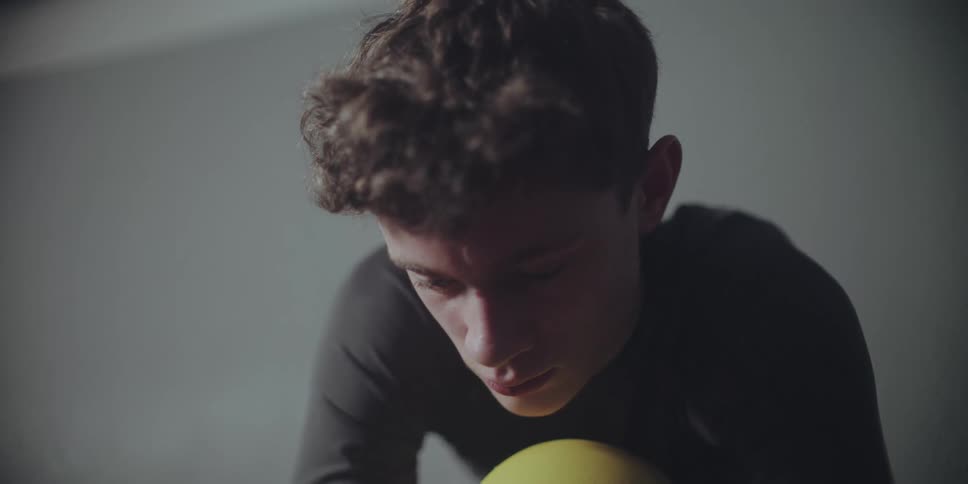 A focused young man with curly brown hair rests his head on a yellow ball during an intense workout