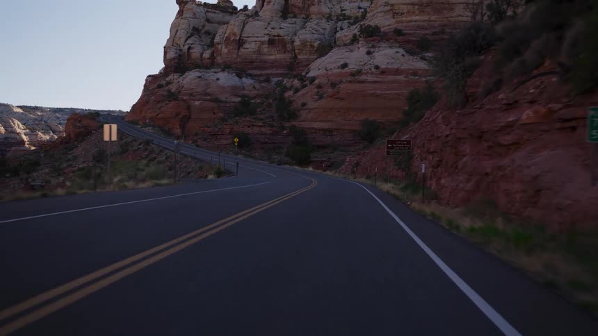 A first-person point-of-view drive along a winding asphalt road through a desert canyon with red rock formations at sunrise