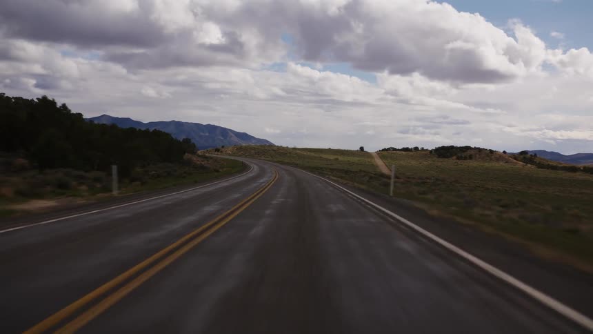 A fast-paced point-of-view drive along a winding two-lane highway through a vast, open landscape with mountains in the distance under a cloudy sky