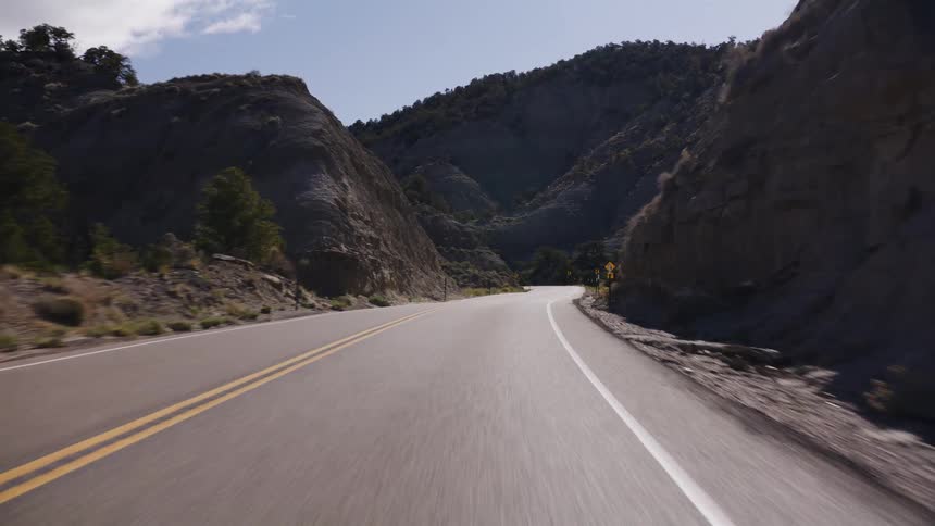 A POV driving shot on a winding paved road through a rugged, arid mountain canyon on a sunny day