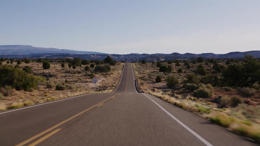 A point-of-view perspective driving along a long, straight, and remote desert road under a clear blue sky