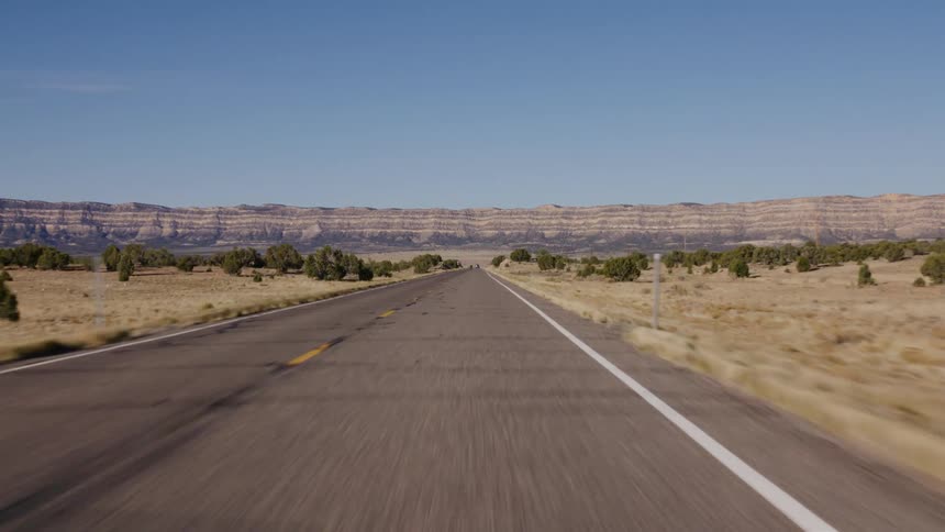 POV driving down a straight desert highway towards distant rock formations on a sunny day